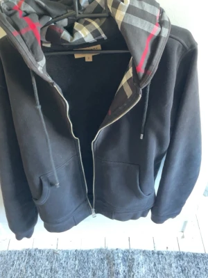Svart hoodie från Burberry med dragkedja - Svart hoodie från Burberry i storlek M med klassiskt rutmönstrat foder i huvan. Tröjan har dragkedja framtill, två fickor och justerbar huva med snören. Perfekt för en stilren och lyxig streetlook.