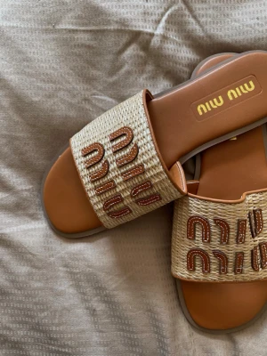 Beige flätade sandaler  - Snygga beige sandaler med flätad ovandel och bruna broderade detaljer. Slip-in modell med öppen tå och platt sula. Perfekta för sommaren och ger en trendig touch till din outfit.