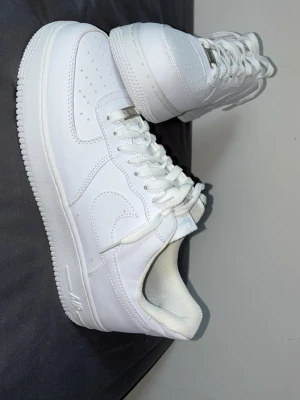 Nike Air Force 1 vita sneakers - Klassiska helvita Nike Air Force 1 sneakers i läder med perforerad tå, vit sula och snörning. Ikonisk Swoosh-logga på sidan och metallbricka med 'AF1' på snörningen. Perfekt för en clean och stilren look.