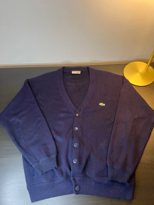 Mörkblå kofta från Lacoste - Snygg mörkblå kofta från Lacoste med klassisk v-ringning och knäppning framtill. Ikoniska gröna krokodilloggan på bröstet. Mjuk och tunnstickad, perfekt för lager-på-lager. Ribbstickade muddar vid ärmslut och nederkant.