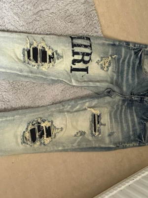 Blå slitna jeans från Amiri - Säljer ett par blå jeans från Amiri med kraftiga slitningar och svarta tygdetaljer under hålen. Jeansen har en cool, tvättad look och tydlig logga på låret. Perfekta för dig som gillar streetstyle och vill sticka ut med din outfit.