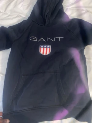 GANT hoodie med broderad logga - Två snygga hoodies från GANT, en i svart och en i vinrött. Båda har broderad GANT-text och sköld med stjärnor och ränder på bröstet. Klassisk huva och stor känguruficka framtill. Perfekt för en avslappnad och sportig stil.