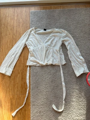 Vit omlottopp med knytband - Snygg vit omlottopp med långa ärmar och v-ringning. Toppen har knytband i midjan som ger en fin passform och en lätt croppad look. Perfekt att styla med jeans eller kjol för en trendig outfit. Har två hål men inget som märks när man har på sig den