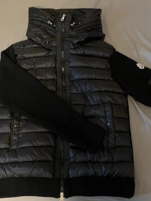 Svart dunväst från Moncler med huva - Snygg svart cardigan från Moncler med quiltad design och huva. Västen har dragkedja framtill, två fickor med knappstängning och Moncler-logga på ärmen. Materialet är en mix av dun och polyester, perfekt för kyliga dagar. Cardiganen är perkfekt till våren och sommaren. Om du är intresserad eller vill ha fler bilder är det bara att skriva.