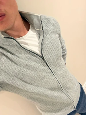 Ljusgrå ziptröja med strukturmönster - Snygg ljusgrå ziptröja med diskret strukturmönster och ståkrage. Tröjan har hel dragkedja framtill och ribbade muddar vid ärmslut och nederkant. Perfekt för lager-på-lager och enkel att matcha med jeans eller chinos.
