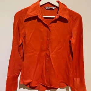 Orange långärmad blus från Zara - Snygg orange blus från Zara med klassisk krage och knäppning framtill. Blusen har långa ärmar och är tillverkad i ett glansigt material som ger en stilren look. Perfekt att styla med jeans eller kjol för en trendig outfit.