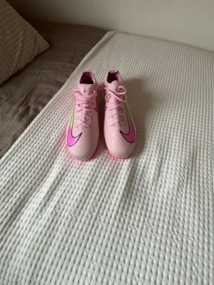 Nike rosa fotbollsskor med neon detaljer - Säljer ett par snygga rosa Nike fotbollsskor med stora svarta och rosa swoosh-loggor samt neongröna detaljer. Skorna har snörning och är tillverkade i syntetmaterial för bra grepp och känsla på planen. Perfekta för dig som vill sticka ut på fotbollsplanen!