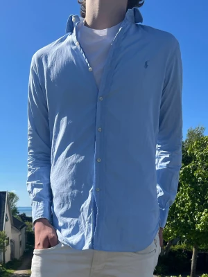 Ljusblå skjorta från Ralph Lauren - (MODDELEN ÄR 185 OCH VÄGER 75kg)(Liten slitning på nacken men stans knappt)Klassisk ljusblå skjorta från Ralph Lauren med button-down krage och broderad logga på bröstet. Skjortan har långa ärmar, vita knappar och är tillverkad i mjuk bomull. Perfekt för en clean och stilren look.