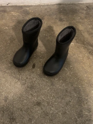 Svarta fodrade gummistövlar - Säljer ett par svarta gummistövlar med mjukt grått foder på insidan. Stövlarna har en klassisk rundad tå och reflexdetalj baktill för extra synlighet. Perfekta för regniga dagar och håller fötterna torra och varma.