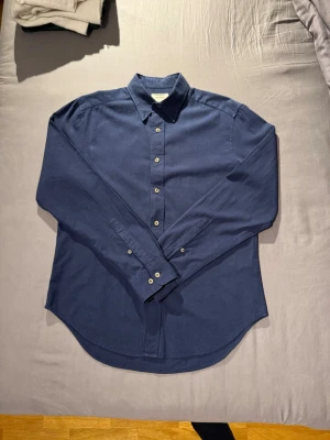 Mörkblå skjorta från EAST WEST   - Snygg blå skjorta från EAST WEST med klassisk krage. Skjortan är långärmad och har en stilren, tidlös design som passar perfekt till många olika outfits. Skjortan har en mycket subtilt randigt mönster. Tillverkad i bomull för en skön känsla och bra passform. Har som ”slits” på sidorna (se bilder). Storlek: M. 