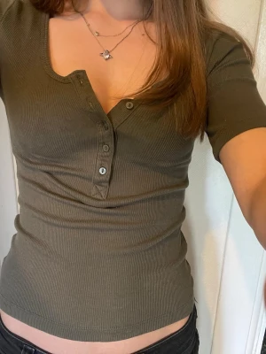 Olivgrön ribbad topp med knappar - Snygg olivgrön ribbad topp med korta ärmar och knappdetalj framtill. Toppen har en v-ringad halsringning och figurnära passform. Perfekt att matcha med jeans eller kjol för en trendig look.
