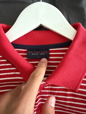 Rödvit randig piké från U.S. Polo Assn - Säljer en röd och vit randig pikétröja från U.S. Polo Assn med klassisk krage och knappar framtill. Tröjan har korta ärmar och är tillverkad i mjuk bomull. Perfekt för dig som gillar sportig och avslappnad stil.