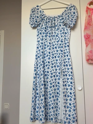 Vit långklänning med blåa blommor - Romantisk långklänning i vit med blått blommönster och puffärmar. Klänningen har en smickrande offshoulder-ringning och är tillverkad i ett lätt, svalt material som passar perfekt för varmare dagar. Snygg slits framtill för extra rörelsefrihet.