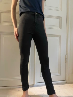 Svarta skinny jeans med hög midja - Säljer ett par svarta skinny jeans med hög midja och klassisk femficksdesign. Jeansen har smal passform hela vägen ner och stängs med knapp och dragkedja. Perfekta för dig som gillar en tight och stilren look.