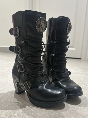 New Rock svarta stövletter med klack - Säljer ett par svarta stövletter från New Rock med  klack , snörning framtill och flera dekorativa spännen i silver. Har använts få gånger men lädret har spruckit på några ställen (se bilder) pga felaktig behandling hos skomakare. 