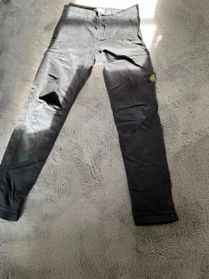 Svarta cargobyxor från Stone Island - Säljer ett par svarta cargobyxor från Stone Island med klassisk patch på benet. Byxorna har smal passform, flera fickor och är tillverkade i ett slitstarkt bomullsmaterial. Perfekta för dig som gillar streetwear och vill ha en clean look. Strl 12 år 156cm men passar längre.