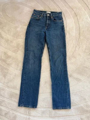 Blå raka jeans med hög midja - Klassiska blå jeans med rak passform och hög midja. Jeansen har fem fickor, knappgylf och subtila slitningar för en avslappnad look. Perfekta till vardags och lätta att matcha med olika stilar.