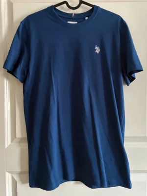 Blå t-shirt med liten logga - Snygg marinblå t-shirt med rund hals och liten vit logga på bröstet. Klassisk passform och tillverkad i mjuk bomull som känns skön mot huden. Perfekt till jeans eller shorts för en clean look.