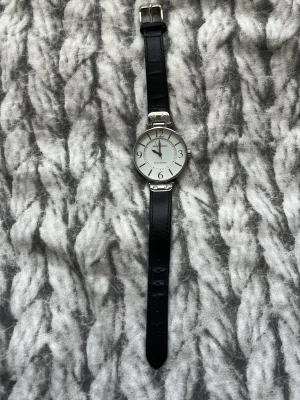 Svart klocka från Anne Klein - Stilren klocka från Anne Klein med svart läderband och silverfärgat runt urtavlan. Urtavlan är vit med tydliga siffror och minimalistisk design. Perfekt för dig som gillar klassiska accessoarer med enkel look.