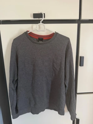 Grå sweatshirt från BOSS - Säljer en stilren mörkgrå sweatshirt från BOSS med diskret broderad logga på bröstet. Tröjan har rund halsringning, långa ärmar och ribbade muddar. Perfekt för dig som gillar enkel och klassisk design. Materialet känns mjukt och bekvämt.