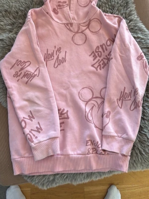 Rosa hoodie med Disney-tryck - Säljer en ljusrosa hoodie med huva och ribbade muddar. Hoodien har tryck med Mickey Mouse och text som 'You're Cool' och 'Enjoy & Relax' i en mörkare rosa nyans. Perfekt för dig som gillar Disney och vill ha en unik look.