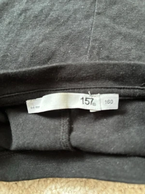 Svart t-shirt från 157 - En enkel svart t-shirt från märket 157. T-shirten har klassisk passform med korta ärmar och rund hals. Materialet är mjukt och bekvämt, perfekt för att matcha med jeans eller shorts. Märkets logga finns på etiketten i nacken.