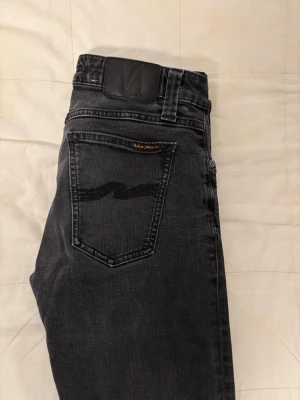 Svarta jeans från Nudie Jeans - Säljer ett par svarta lite mer slimmade jeans från Nudie Jeans. Jeansen har klassisk femficksdesign, vågdetalj på bakfickorna och är gjorda i mjukt denimtyg. Perfekta för dig som gillar en clean och modern look. Storlek W30 L32. Priset är inte hugget i sten. Tveka inte att höra av er vid minsta fråga.