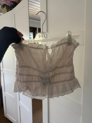 Ljus volang topp med snörning - Säljer en ljus, croppad topp med volangkant och snörning framtill. Toppen är ärmlös och har ett lätt transparent material med rynkade detaljer som ger en cool och romantisk vibe. Perfekt för dig som gillar trendiga och feminina plagg. Prislappen kvar, aldrig använt, storleken är i M men skulle säga att den passar mer mig som är en S