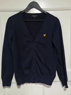 Mörkblå kofta från Lyle & Scott - Snygg mörkblå kofta från Lyle & Scott med klassisk gul logotyp på bröstet. Koftan har v-ringning, långa ärmar och knäppning med svarta knappar framtill. Perfekt för lager-på-lager och stilrena outfits.