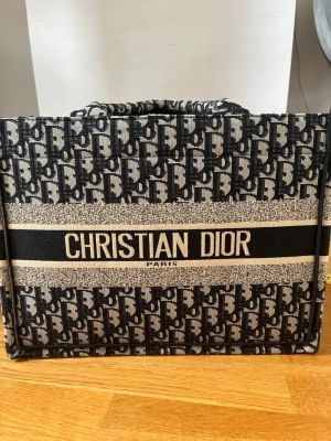 Christian Dior Book Tote shoppingväska - Säljer en ikonisk Book Tote shoppingväska från Christian Dior i svart och beige canvas med Dior-logga över hela väskan. Väskan har rektangulär form och breda handtag, perfekt för att bära allt du behöver. Märke och ursprung: Christian Dior, Paris, Made in Italy.
