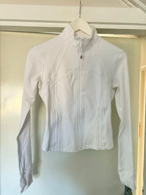 Vit träningsjacka från Lululemon - Snygg vit träningsjacka från Lululemon med figurnära passform och dragkedja framtill. Använd en gång! 