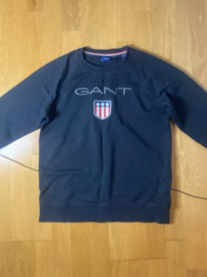 Mörkblå sweatshirt från GANT - Snygg mörkblå sweatshirt från GANT med tryckt logga och sköld på bröstet. Tröjan har rund halsringning, långa ärmar och ribbade muddar. Perfekt för en avslappnad och stilren look.