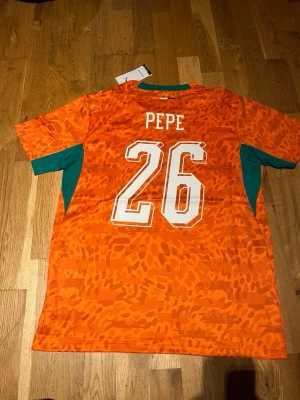 Elfenbenskusten matchtröja Puma orange - Snygg orange matchtröja från Elfenbenskustens landslag med nummer 26 och namnet Pepe på ryggen. Tröjan har gröna detaljer på sidorna och ärmarna, samt vita tryck. Tillverkad i lätt och ventilerande material, perfekt för fotboll eller träning.