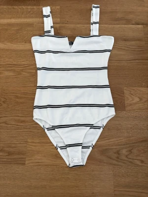 Vit body med svarta ränder från Bershka - Snygg vit body från Bershka med svarta horisontella ränder och breda axelband. Materialet är mjukt och stretchigt, perfekt för att skapa en clean och modern look. Praktisk knäppning i grenen och cool V-ringning framtill.