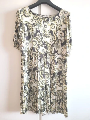 Odd Molly Klänning med paisleymönster - Odd Molly klänning med ett mönster i svart, vitt, gult och grönt. Klänningen har korta ärmar med volangdetaljer och en rund halsringning. Midjan är markerad med en resår som skapar en smickrande silhuett. Klänningen faller i flera volanger ner till knäna.