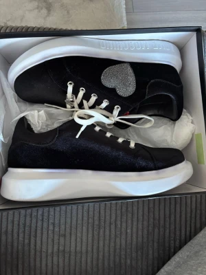 Love Moschino svarta sneakers med hjärta - Svarta sneakers från Love Moschino med vit sula och vita snören. På sidan finns ett glittrigt silverhjärta som detalj. Skorna har en låg profil och är tillverkade i ett material som ser ut som sammet. Perfekt för dig som vill sticka ut med coola detaljer.