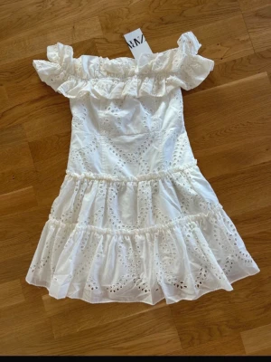 Vit offshoulder klänning från Zara - Söt vit kortklänning från Zara med offshoulder och volangdetaljer. Klänningen har broderade spetsmönster och är tillverkad i bomull. Perfekt för sommarens alla tillfällen och ger en romantisk vibe.
