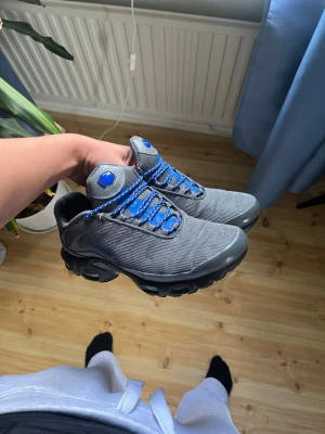 Nike Air Max Plus grå/blå sneakers - Nike Air Max Plus sneakers i grå mesh med blå detaljer och svarta sulor. Skorna har blå snören och ikoniska Air Max-bubblor i sulan för extra komfort. Perfekt för dig som vill ha en cool och sportig look.