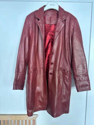 Vinröd läderjacka, 36 - Säljer en lång vinröd läderjacka med klassisk krage, knäppning framtill och två fickor. Jackan har en snygg, rak passform och är fodrad med matchande vinrött tyg. Storleken går mer åt 38. 