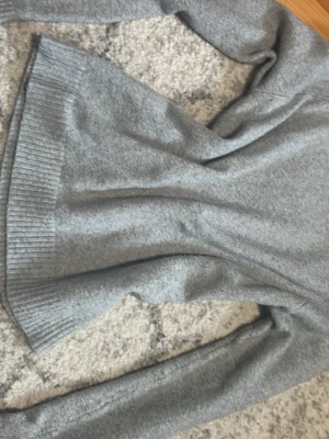 Grå stickad tröja med ribbad kant - En enkel och stilren grå stickad tröja med långa ärmar och ribbad nederkant. Perfekt för lager-på-lager och passar till jeans eller kjol. Materialet är mjukt och bekvämt, vilket gör den skön att ha på sig hela dagen.