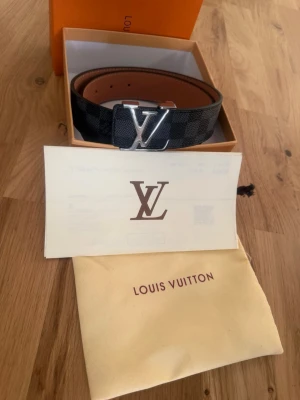 Louis Vuitton svart rutigt bälte - Säljer ett exklusivt svart bälte från Louis Vuitton med rutigt mönster och en stor silverfärgad LV-logga på spännet. Bältet är tillverkat i skinn och kommer med originalbox och dustbag. Perfekt för att lyfta din outfit med en lyxig touch.