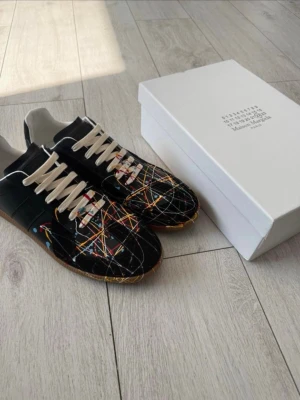 Maison Margiela svarta sneakers - Maison Margiela sneakers i svart med färgglada stänk och vita skosnören. Skorna har en låg silhuett och är tillverkade i en mix av mocka och läder. Unik design med färgade linjer över hela ovandelen för en kreativ vibe.