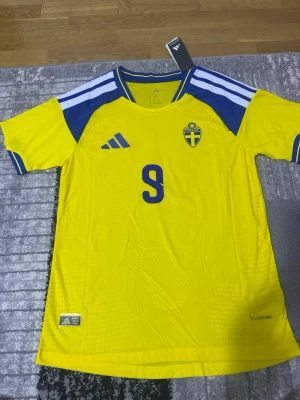Svensk landslagströja Adidas gul - Säljer en gul svensk landslagströja från Adidas med blå detaljer och nummer 9 på bröstet. Tröjan har Sveriges emblem på vänster sida och klassiska Adidas-ränder på axlarna. Materialet är lätt och andas, perfekt för fotboll eller träning. Player version 