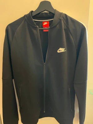 Svart zip hoodie från Nike - Säljer en svart zip hoodie från Nike med vit logga på bröstet. Tröjan har långa ärmar, dragkedja framtill och två fickor. Materialet är mjukt och sportigt, perfekt för en avslappnad och cool look.