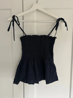 Marinblå linnetopp med knytband - Supersöt marinblå linnetopp med smockad överdel och knytbara axelband. Toppen har en luftig och lätt volang nertill.