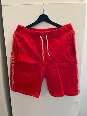 Röda shorts med snörning från Hugo  - Snygga röda shorts från Hugo med vit snörning i midjan och vit logotyp längs sidorna. Shortsens material är mjukt och bekvämt, perfekt för sport eller chill. De har ribbad hög midja och praktiska fickor på sidorna.