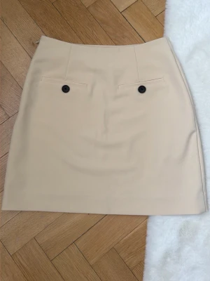 By Malene Birger - Stilren beige kortkjol med två svarta knappar framtill och diskreta fickor. Kjolen har en rak och enkel form som ger ett klassiskt intryck. Endast använd en gång. Nypris 1899kr.