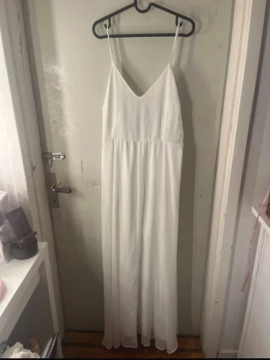 Vit långklänning med knyt i ryggen - Säljer en elegant vit långklänning med smala axelband och v-ringning. Klänningen har ett fint knyt i ryggen som ger en extra detalj. Perfekt för sommarens alla tillfällen. Materialet känns lätt och luftigt. Den har en pytteliten fläck vid v-ringninfdn men det är på insidan av klänningen (se bild 3)