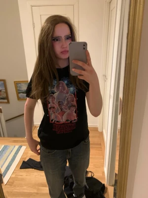 Svart Stranger Things t-shirt - Cool svart t-shirt med Stranger Things-motiv på framsidan. Trycket visar flera karaktärer från serien och har färgglada detaljer i rött och blått. T-shirten har rund hals och korta ärmar, perfekt för fans av serien.