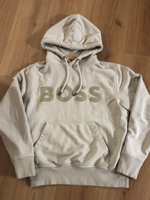 Hoodie Hugo Boss  - Snygg beige hoodie från BOSS med stor logga på bröstet. Hoodien har en klassisk passform, ribbade muddar och en stor känguruficka framtill. Materialet är mjukt och bekvämt, perfekt för chill dagar. Justerbar huva med snören och stilren design.